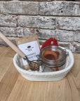 Picture with danish dough whisk, glass jar, packet of sourdough starter and a red silicone bowl scraper inside of a rattan banneton
