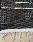 Close-up of a woven rug with a dark charcoal-brown body, off-white horizontal stripe accents, and a fringed edge, laid on a light wood surface.