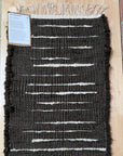 A rectangular woven rug with a dark charcoal-black base and horizontal off-white stripe patterns, displayed flat on a light wood floor next to a brick column. A small product information card is attached to the upper left corner.