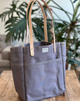 Gray tote bag with brown leather straps on a wooden table with plants in the background
