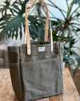 Green tote bag with leather straps on a wooden surface with plants in the background