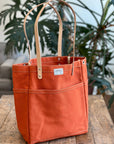 Orange tote bag with leather handles on a wooden table, plant and sofa in the background