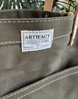Gray tote bag with 'Artifact' label against a blurred green plant background