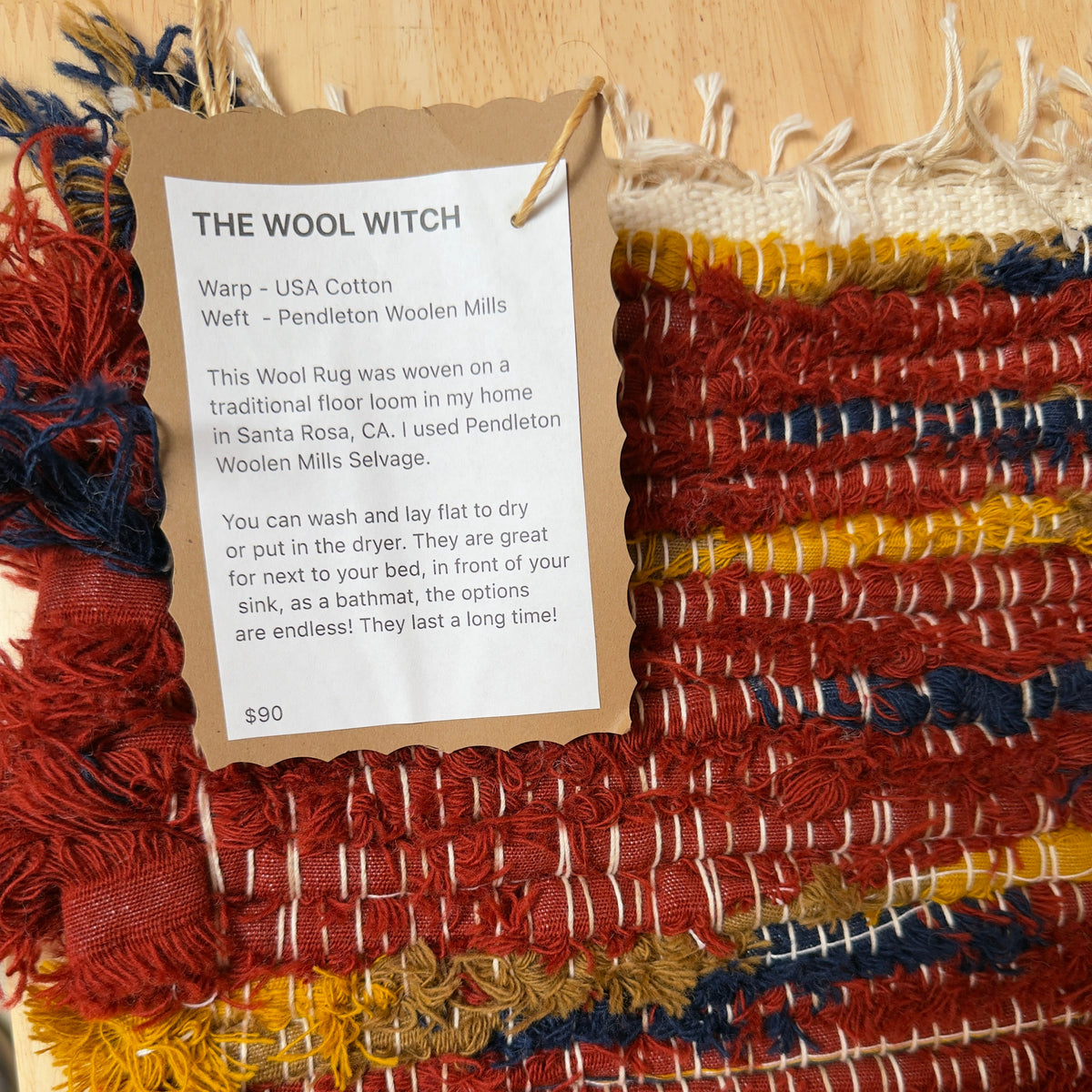 A handmade woven rug with a kraft paper tag reading 'THE WOOL WITCH' and product details, displayed on a light wood surface. The rug features a multicolor pattern of red, navy blue, mustard yellow, and cream.