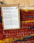 A handmade woven rug with a kraft paper tag reading 'THE WOOL WITCH' and product details, displayed on a light wood surface. The rug features a multicolor pattern of red, navy blue, mustard yellow, and cream.
