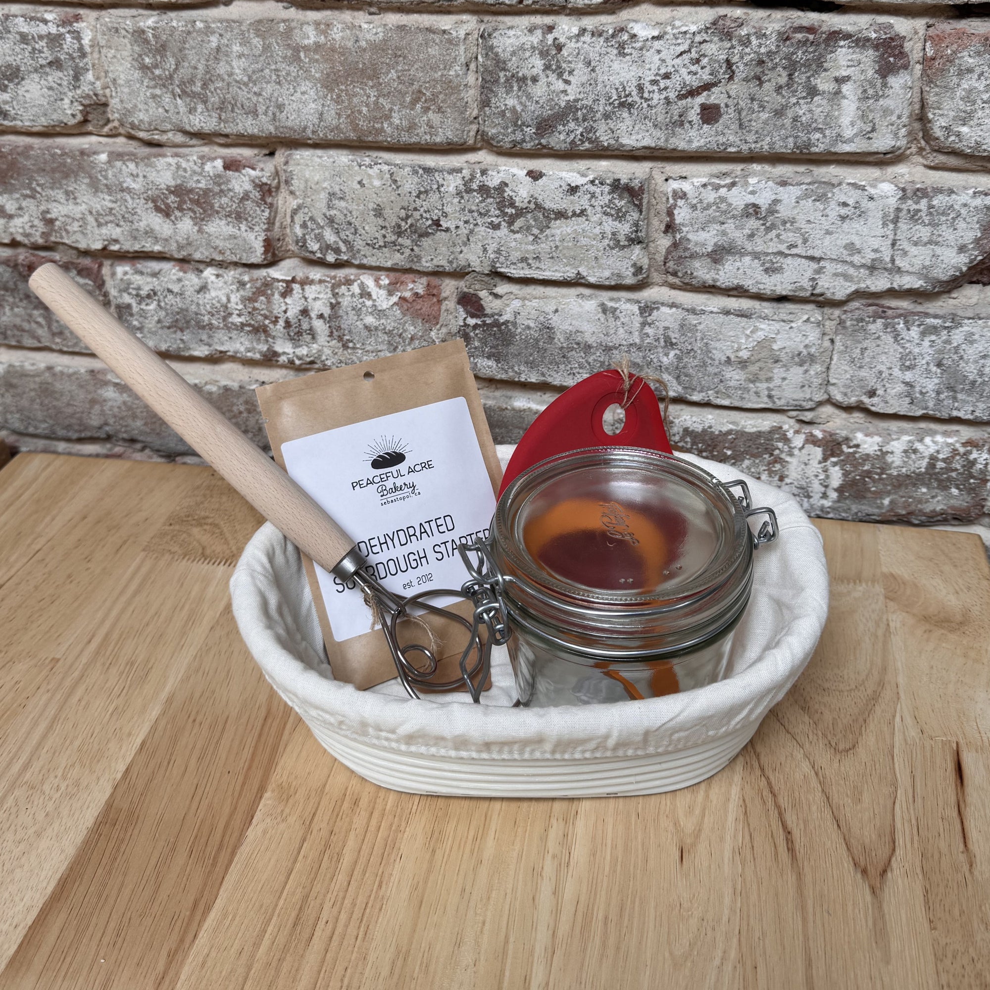 Picture with danish dough whisk, glass jar, packet of sourdough starter and a red silicone bowl scraper inside of a rattan banneton