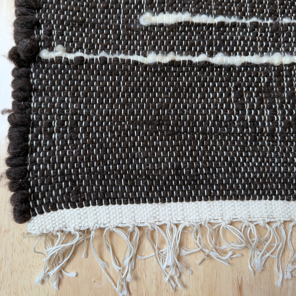 Close-up of a woven rug with a dark charcoal-brown body, off-white horizontal stripe accents, and a fringed edge, laid on a light wood surface.