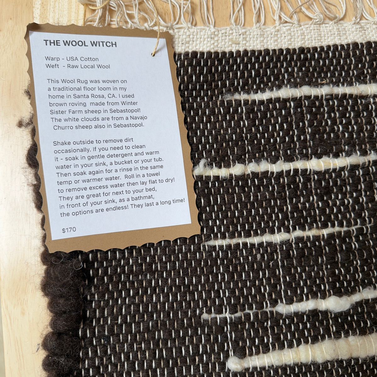 A close-up of a woven wool rug with a black and white striped pattern, showing a white paper tag with product details for 'The Wool Witch' brand.