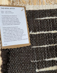 A close-up of a woven wool rug with a black and white striped pattern, showing a white paper tag with product details for 'The Wool Witch' brand.