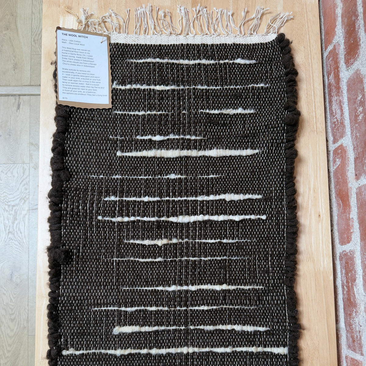 A rectangular woven rug with a dark charcoal-black base and horizontal off-white stripe patterns, displayed flat on a light wood floor next to a brick column. A small product information card is attached to the upper left corner.