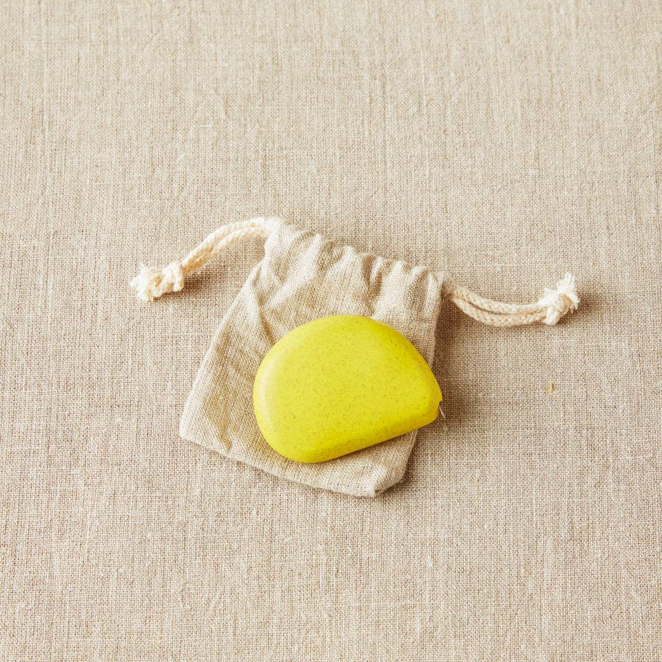 Mustard seed colored Cocoknits tape measure on a white drawstring bag on a beige fabric background