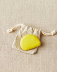 Mustard seed colored Cocoknits tape measure on a white drawstring bag on a beige fabric background